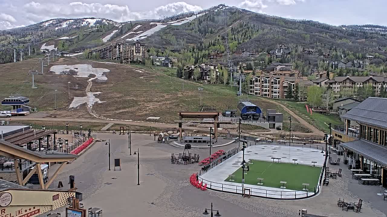 A partially snow-covered mountainside overlooks a plaza with an outdoor rink and seating areas under a partly cloudy sky.