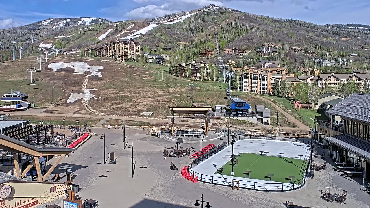 A mostly sunny day reveals a mountainside village with a paved plaza featuring a partially melted ice rink, outdoor seating areas, and ski lifts ascending a snow-dusted mountain.