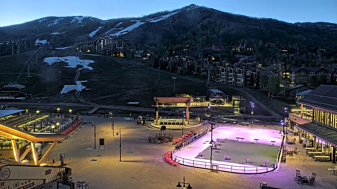 An illuminated ice rink sits in a plaza at the base of a snow-dusted mountain at dusk.