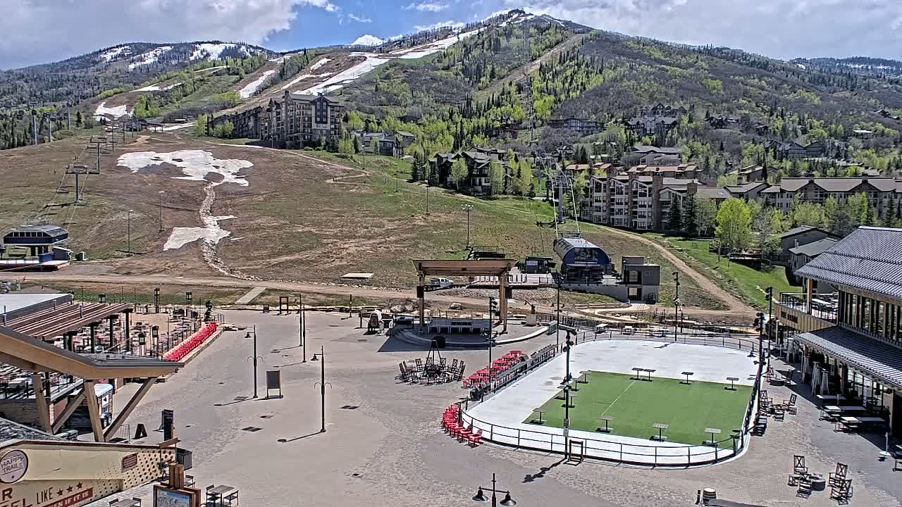 An outdoor plaza with a small artificial turf field, surrounded by buildings and a mountainside with patches of snow under a partly cloudy sky.