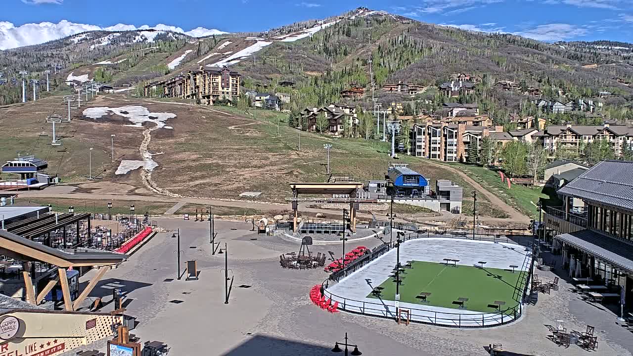 A mountainside village with buildings, ski lifts, and a paved plaza containing a green rink and outdoor seating, all under a sunny sky.