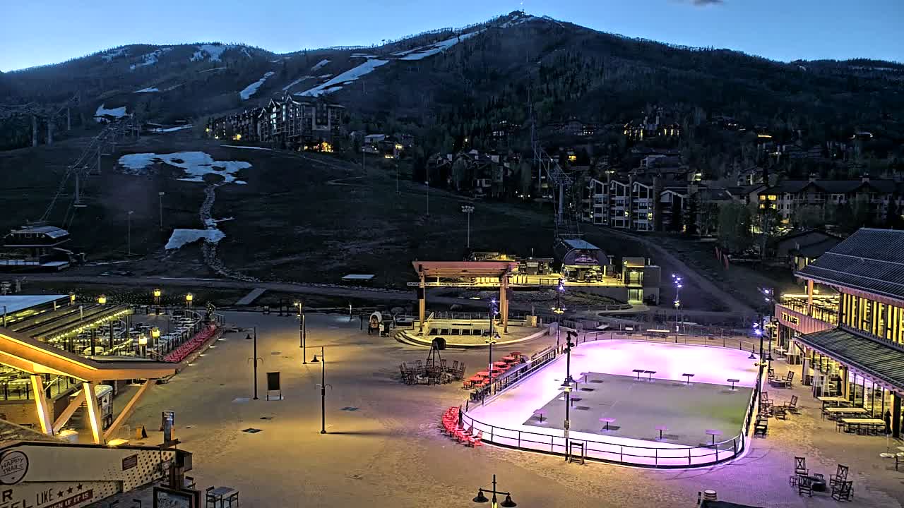 An illuminated outdoor ice rink sits in a mountain village at dusk, with a ski resort visible on a nearby mountain.