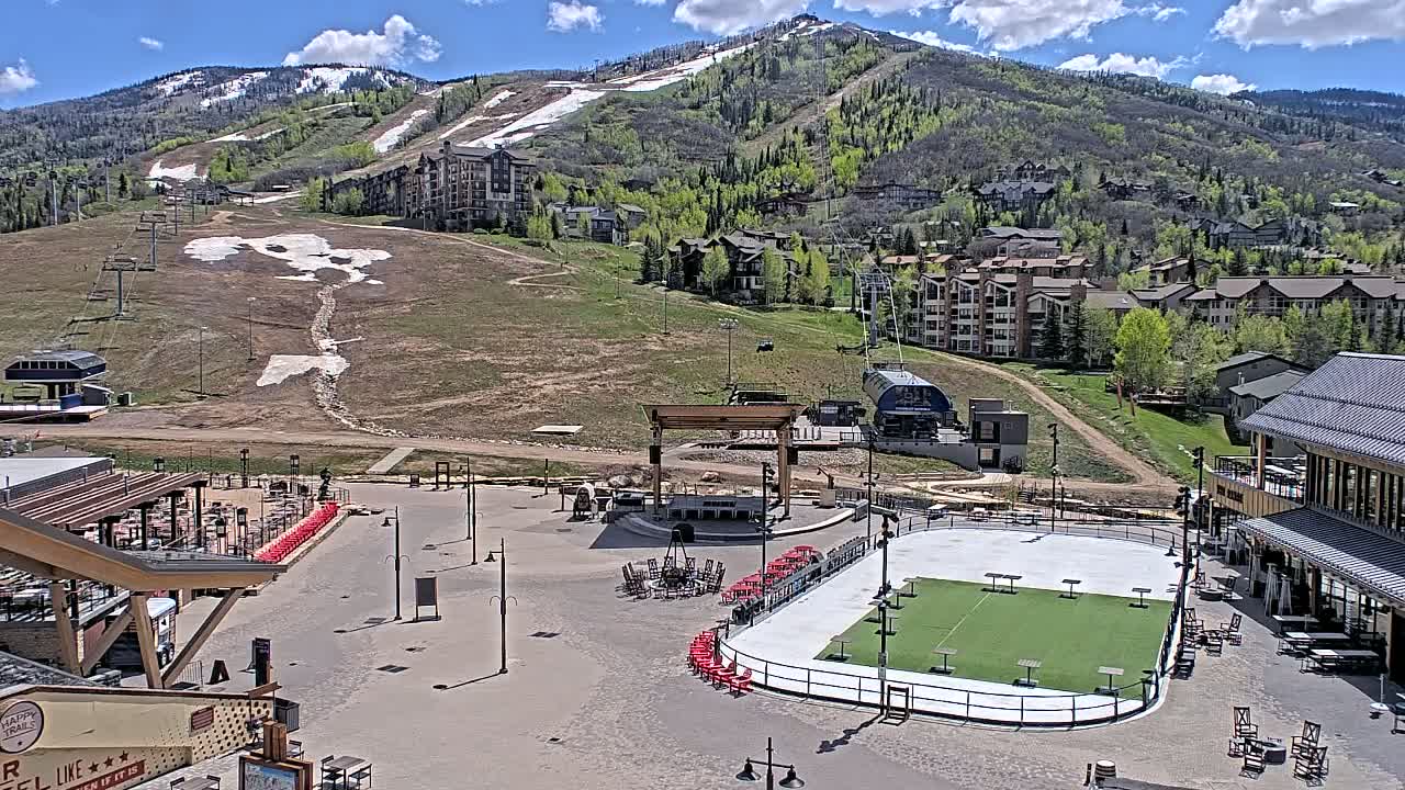 A sunny day reveals a mountain resort town with a paved plaza containing an outdoor rink and stage, backed by a snow-dusted mountain range and ski lift.