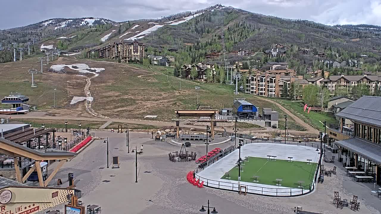 A mountainside village with buildings and ski lifts, under partly cloudy skies, features a paved plaza with an ice rink, outdoor seating, and a stage.
