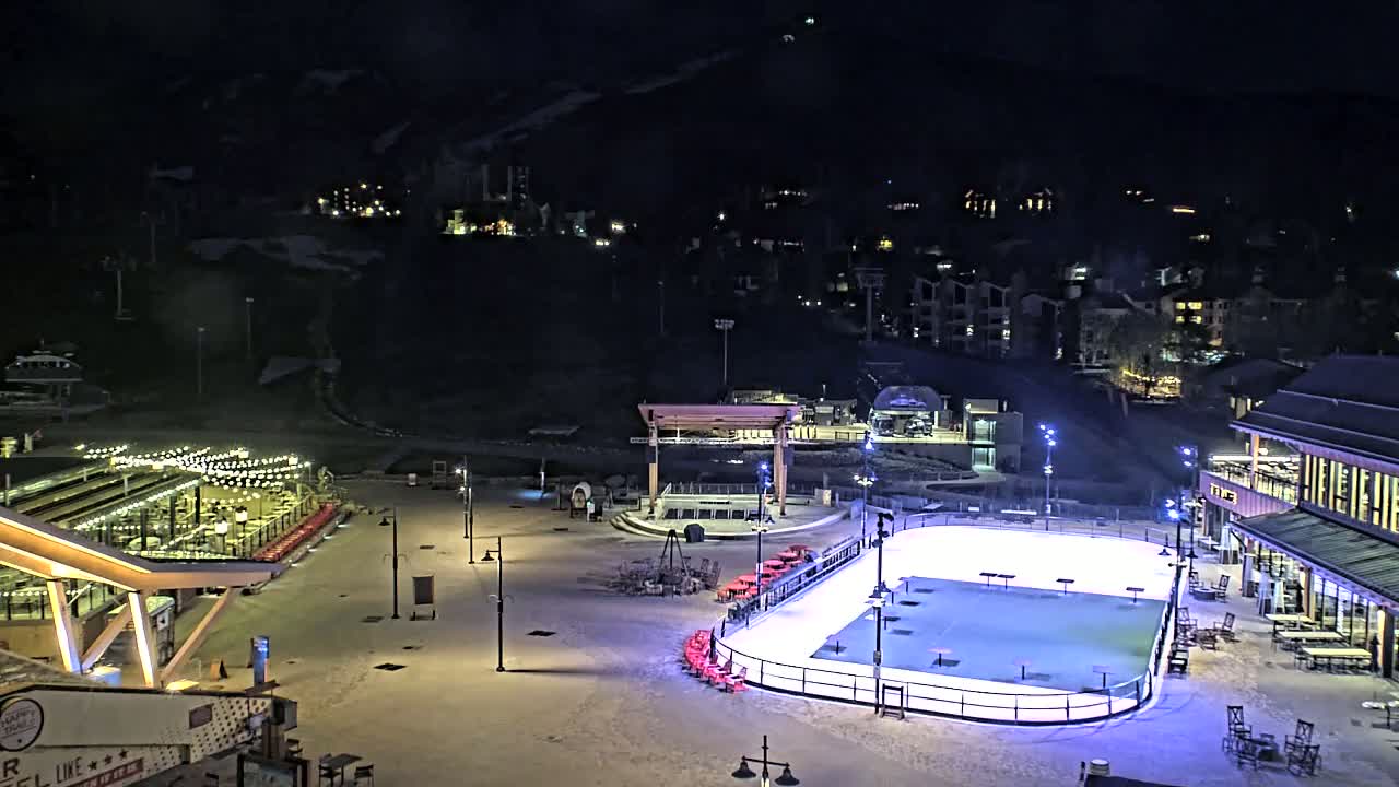 An outdoor ice rink is illuminated at night, surrounded by snow-covered ground, buildings, and outdoor seating areas.