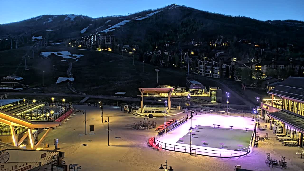 An illuminated ice rink sits in a plaza at the base of a snow-dusted mountain at twilight.