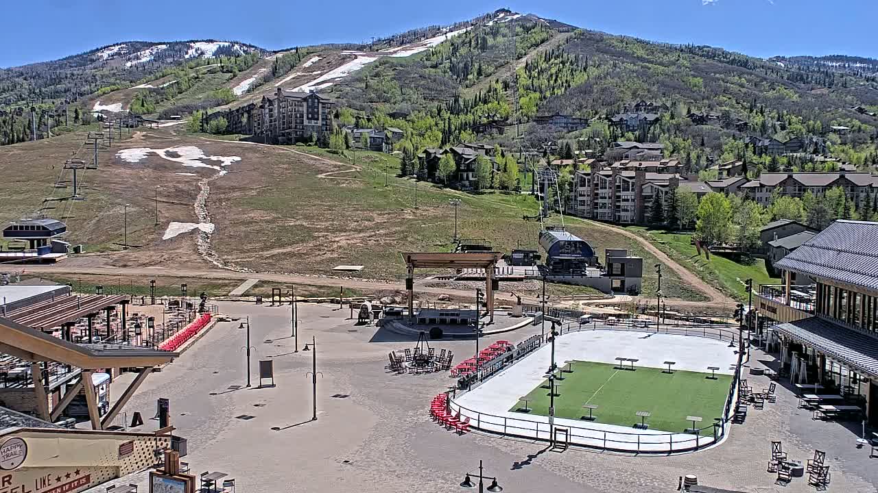 A sunny day reveals a mountain resort village with a paved plaza containing an outdoor rink, a stage, and seating areas, all situated below a mountain with remnants of snow and ski lifts.