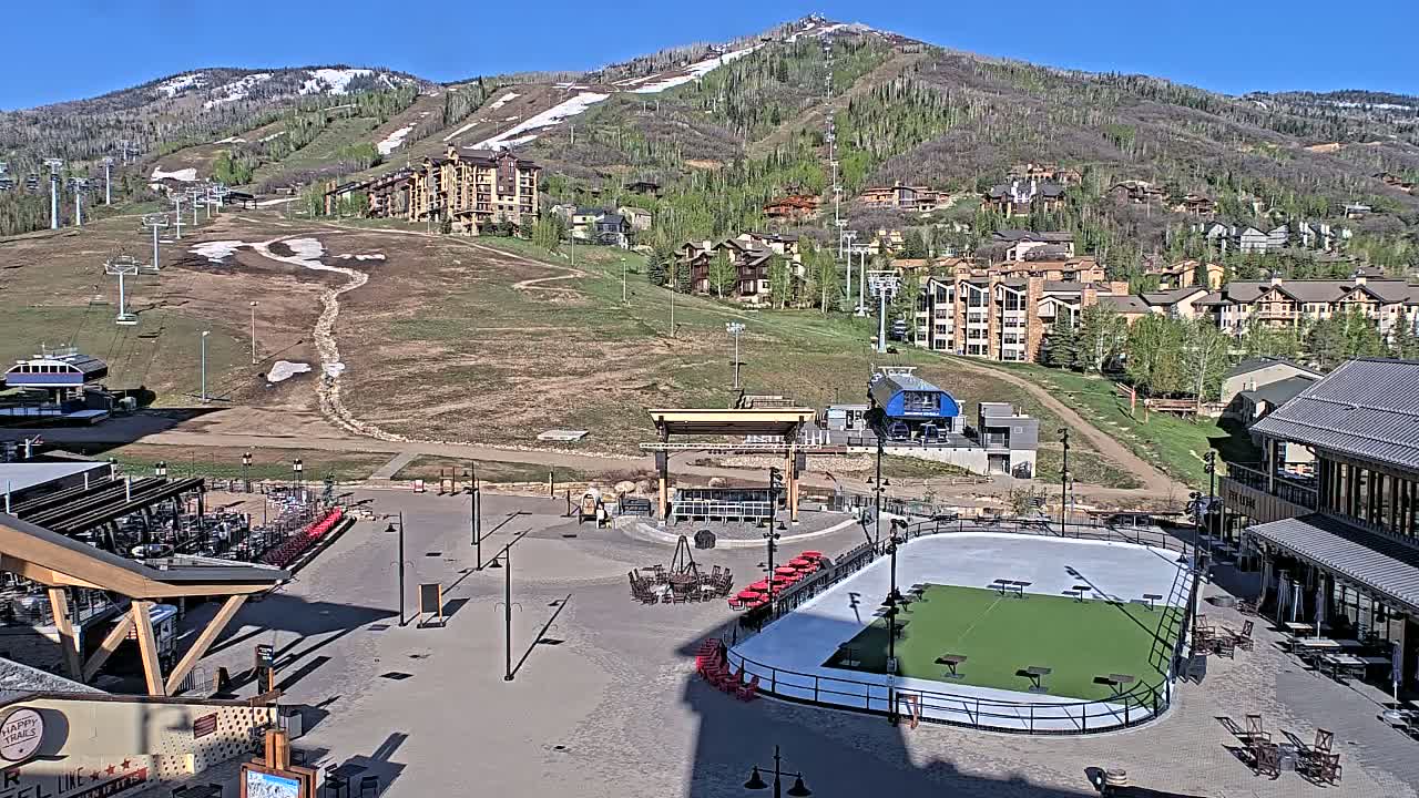 A sunny day reveals a mountainside resort village with a paved plaza featuring an ice rink, outdoor seating, and a stage, all overlooking ski slopes and buildings.