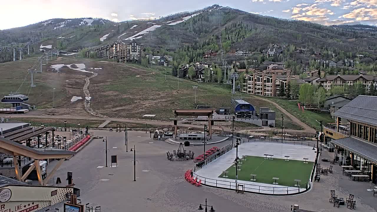A mountainside village with a ski lift and a paved plaza containing an outdoor rink and seating areas is visible under a partly cloudy sky.
