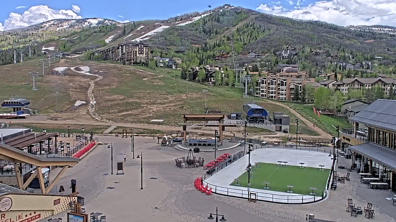 A mostly sunny day reveals a mountainside resort with a paved plaza containing an outdoor rink, seating areas, and buildings, all overlooking a ski slope with remnants of snow.