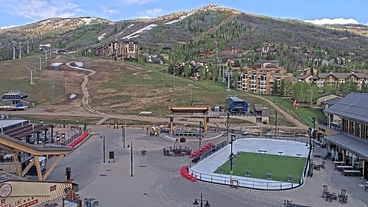 A mostly sunny mountainside view shows a village with buildings, ski lifts, and a paved plaza containing an ice rink, outdoor seating, and a few people.