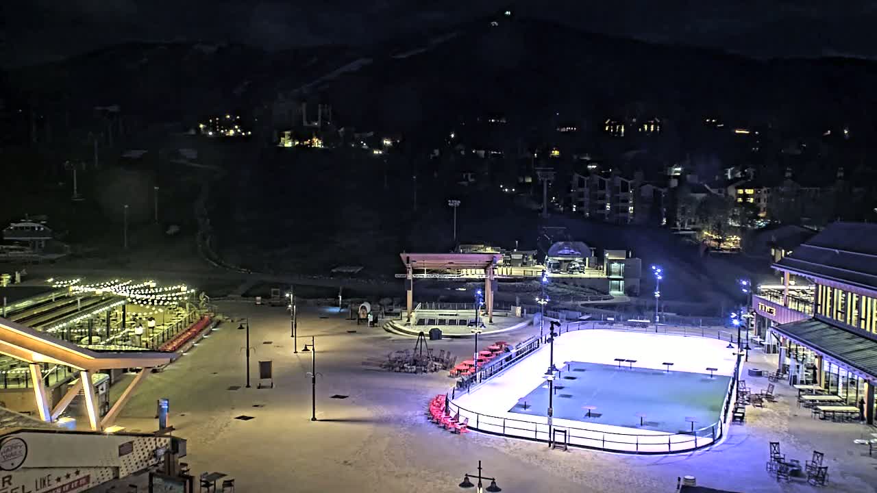 A nighttime, snow-covered plaza features an illuminated ice rink, outdoor seating areas, and buildings nestled against a dark mountainside.