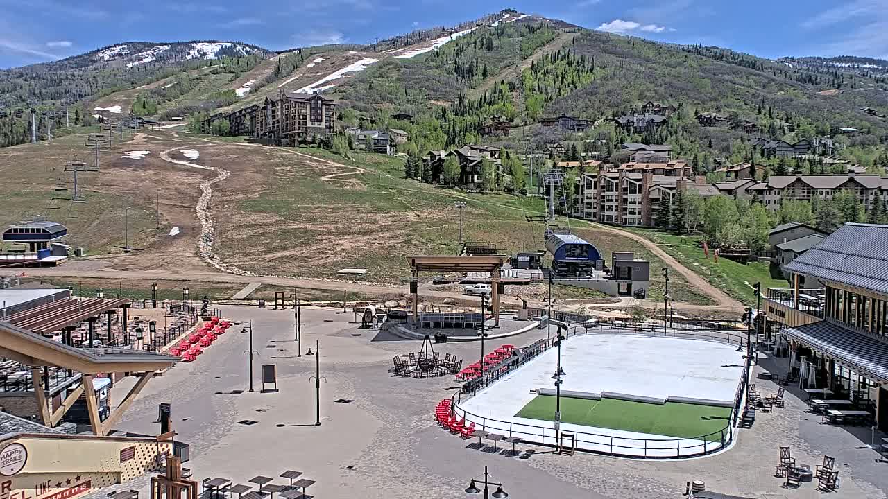 A paved plaza with a rink, outdoor seating, and a stage sits below a ski resort on a sunny day with a mountain in the background.