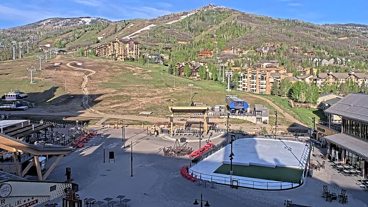 An outdoor scene on a sunny day shows a mountainside with ski lifts and buildings, a plaza with an ice rink and outdoor seating areas.
