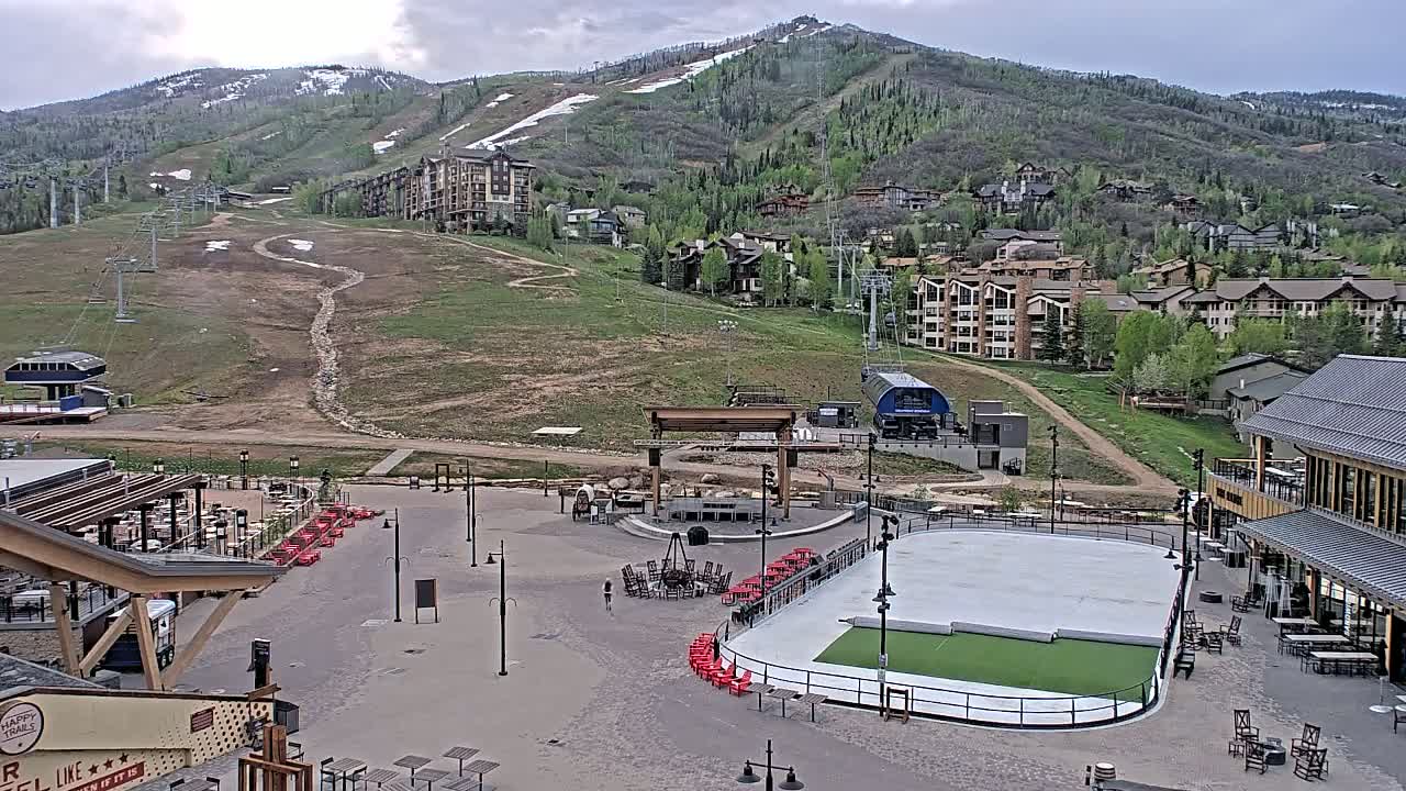 A mostly cloudy day reveals a mountainside village with a ski resort, featuring buildings, chairlifts, a plaza with a covered ice rink, outdoor seating areas, and a fire pit.
