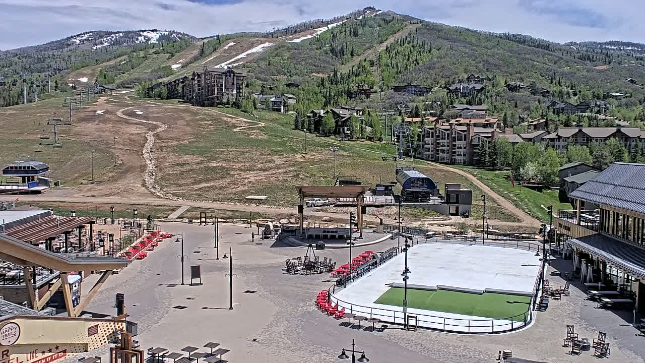 A mountainside village with an empty ice rink and outdoor seating areas is visible on a partly cloudy day.