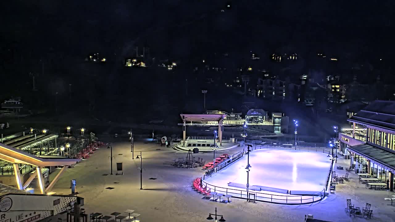 An outdoor ice rink is illuminated at night, surrounded by snow-covered ground, buildings, and seating areas.