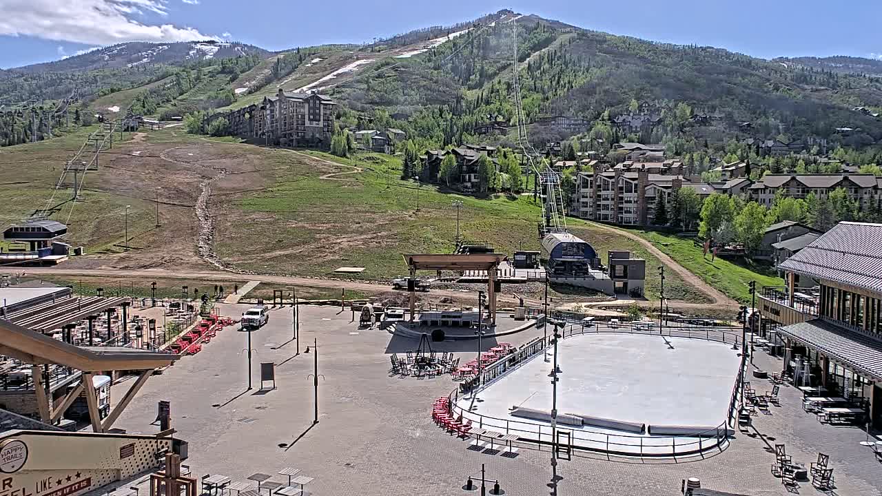 A sunny mountainside village with a paved plaza containing a rink and outdoor seating areas overlooks a ski resort with several chairlifts.