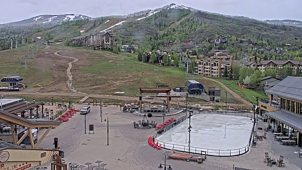 An empty ice rink and outdoor seating area sit below a mountain village on a cloudy day, with a ski lift and mountain slopes visible in the background.