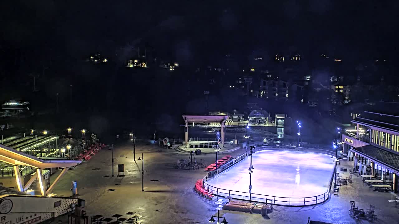An outdoor ice rink is illuminated at night, surrounded by buildings and a plaza with scattered seating, under a dark, possibly cloudy sky.