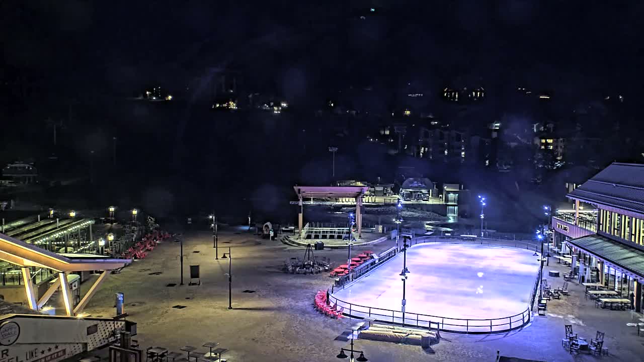 An outdoor ice rink is illuminated at night, surrounded by buildings and sparsely populated snow-covered ground under a dark sky.