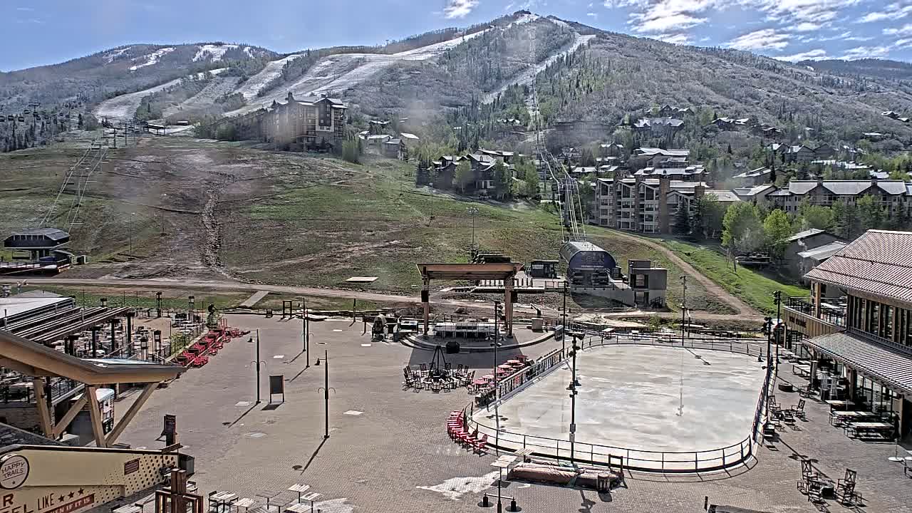 An outdoor plaza with a skating rink and a stage is situated below a mountain with some remaining snow patches, under a mostly sunny sky.