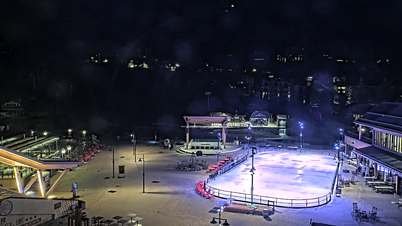 An outdoor ice rink, illuminated at night, is surrounded by buildings and sparsely populated snow-covered ground under a dark, cloudy sky.