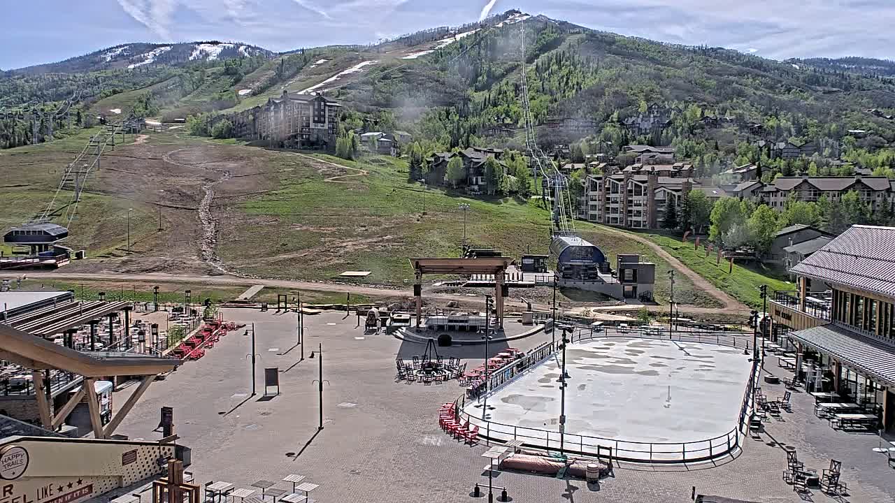 A mostly sunny day reveals a mountainside ski resort with a plaza featuring an empty ice rink, outdoor seating, and a stage, all overlooking a grassy hillside dotted with buildings and ski lifts.