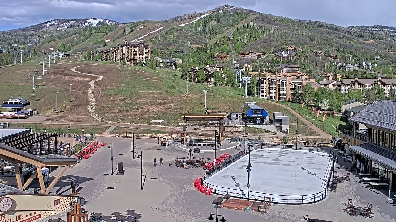 An outdoor plaza with a skating rink, surrounded by buildings and a mountain with ski lifts in the background under partly cloudy skies.