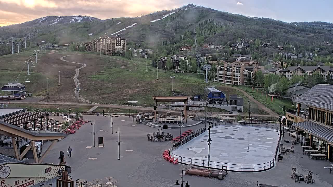 An outdoor scene shows a mostly empty plaza with an ice rink, seating areas, and a stage, situated at the base of a mountain with residential buildings and ski lifts under partly cloudy skies.