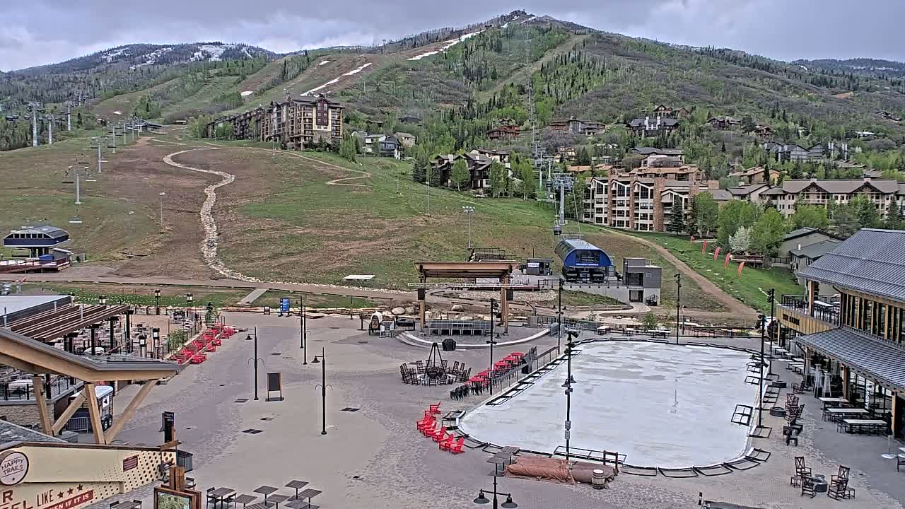 An outdoor scene shows a mostly empty plaza with a covered ice rink, buildings, ski lifts, and a mountainside with green vegetation under a cloudy sky.
