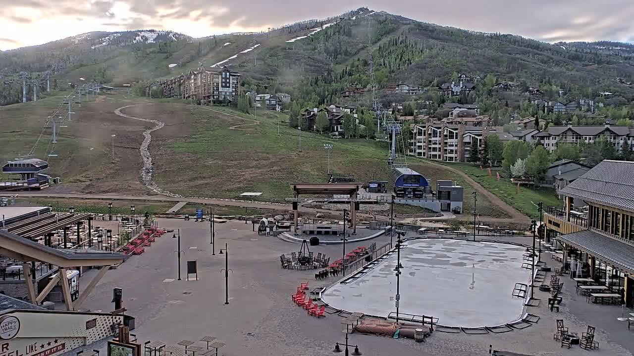 An outdoor plaza with a skating rink, outdoor seating areas, and ski lifts visible on a mountainside in the background under a partly cloudy sky.