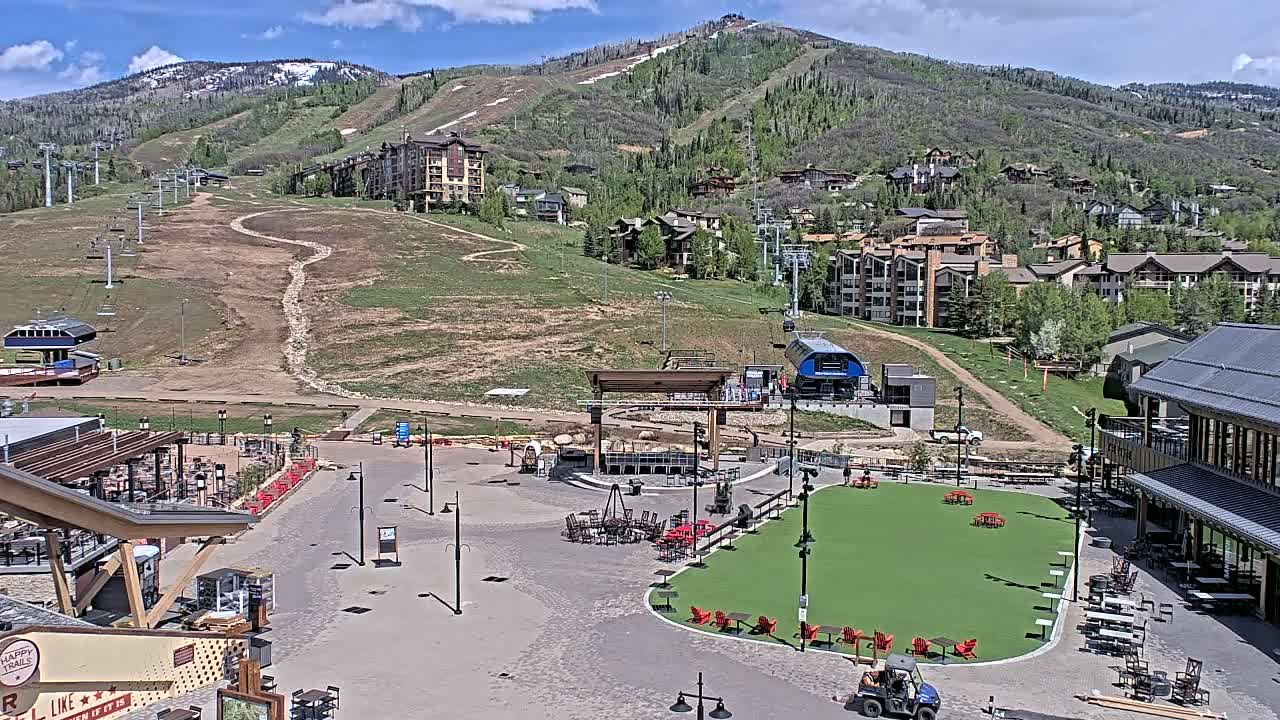 A mountainside village with ski lifts, buildings, and a plaza featuring a grassy area with outdoor seating, all under sunny skies.