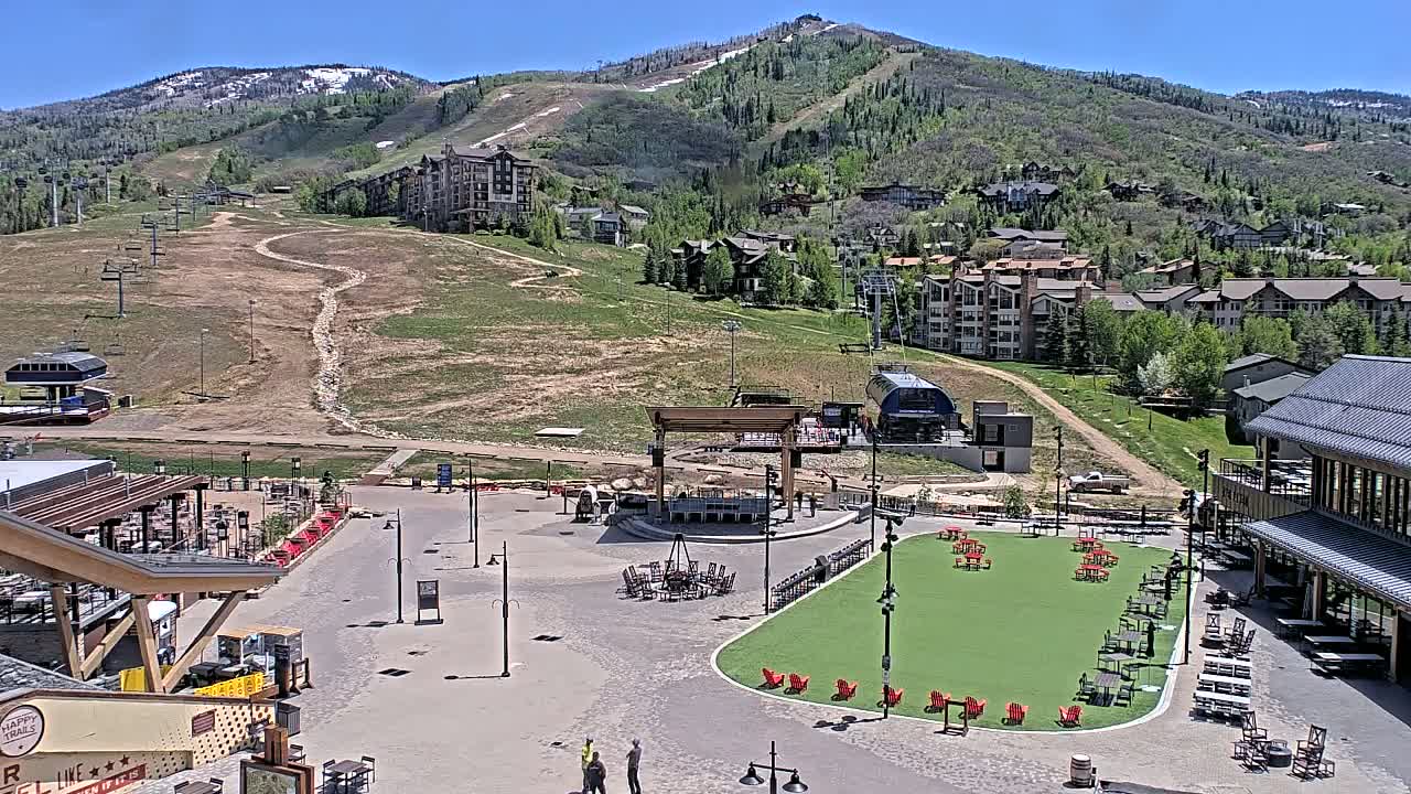A sunny mountainside village square with a green lawn, outdoor seating, and buildings, overlooks a grassy hillside with ski lifts and slopes.
