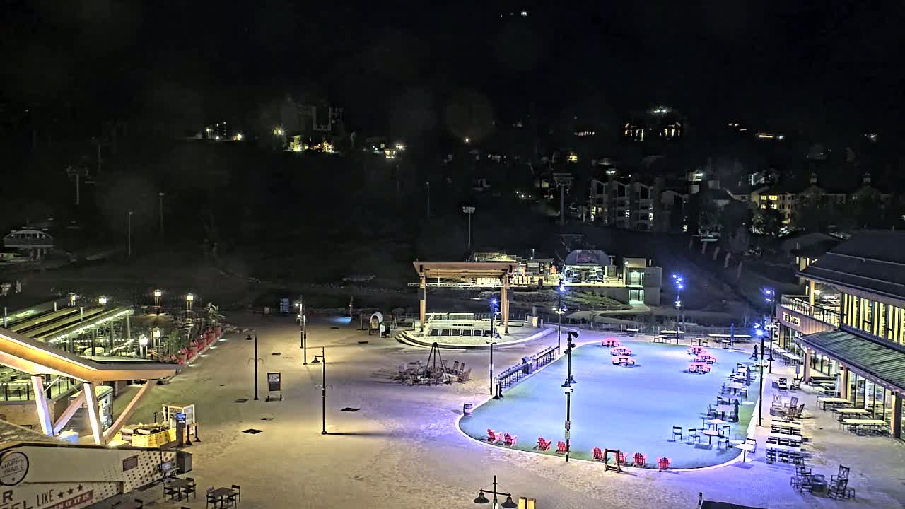 A snow-covered plaza at night with illuminated buildings, outdoor seating, and a rink-like area in the center.