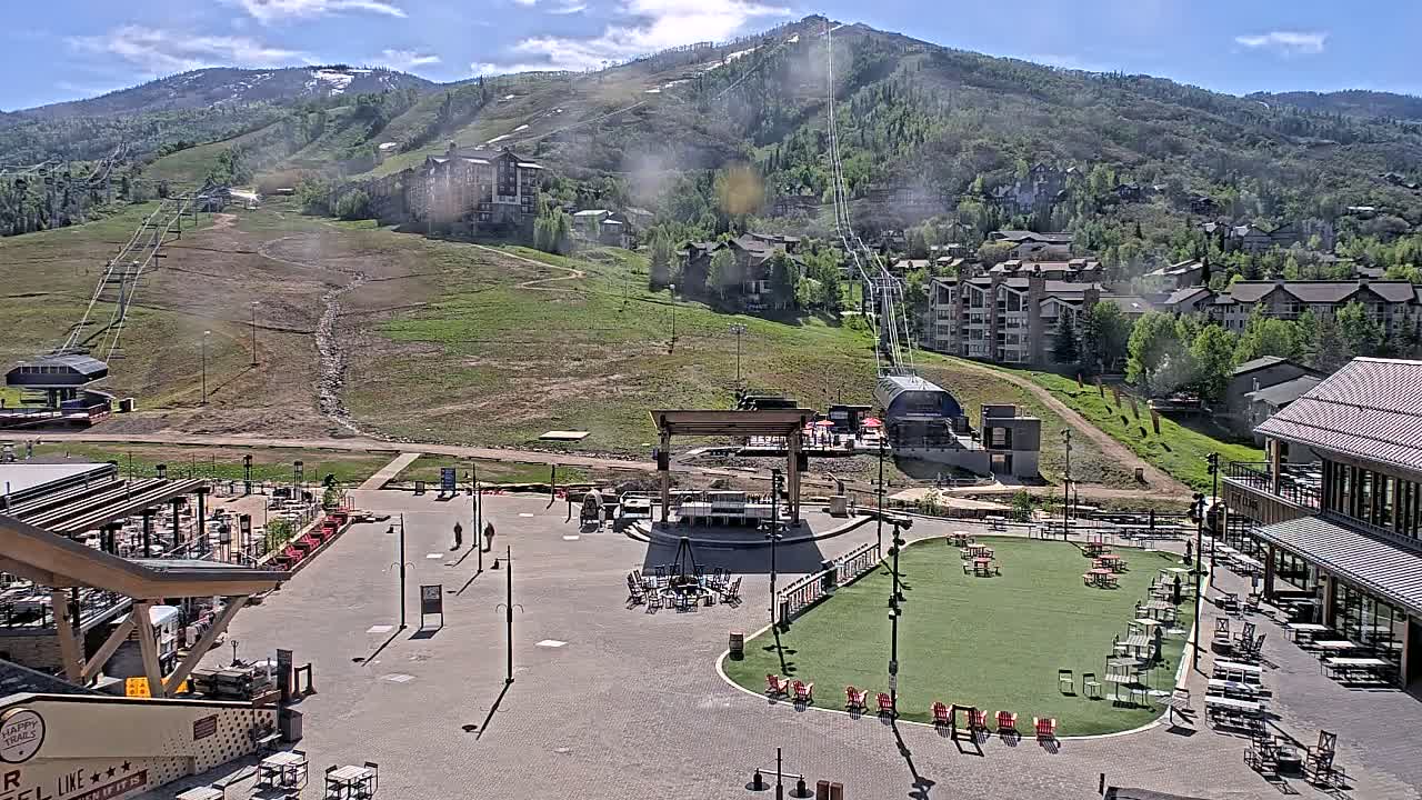 A mountainside village square with a grassy area, outdoor seating, and a stage sits below a ski lift ascending a mountain on a sunny day.