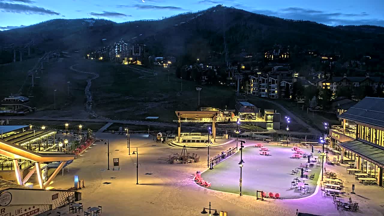 An illuminated plaza with buildings and a stage sits below a dark mountain at dusk, with ski lifts visible on the mountainside.