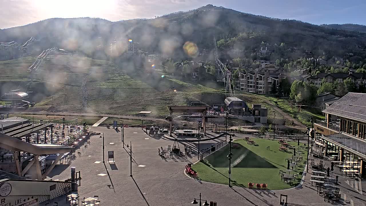 A hazy, sunny day reveals a mountainside ski resort base area with a plaza containing outdoor seating areas, a stage, and a grassy lawn, all situated below a mountain with ski lifts visible.