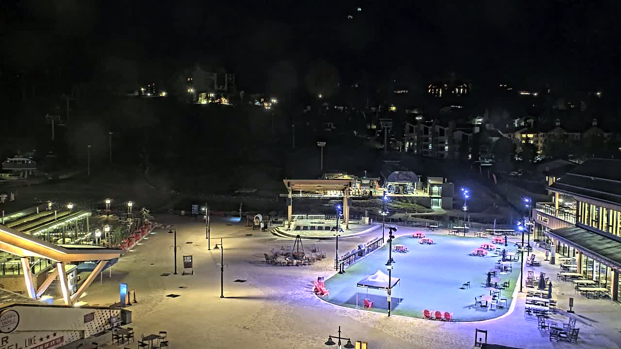 A nighttime view of a snow-covered plaza with outdoor seating, a stage, and buildings, illuminated by artificial light, with a hill of buildings in the background under a clear, dark sky.