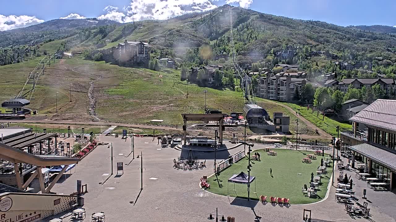 A sunny mountain resort base area with a paved plaza, a grassy area with outdoor seating, a stage, and chairlifts ascending a mountainside.