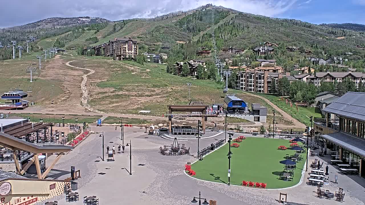A mountainside village with ski lifts, buildings, and a paved plaza featuring a green space and outdoor seating under partly cloudy skies.