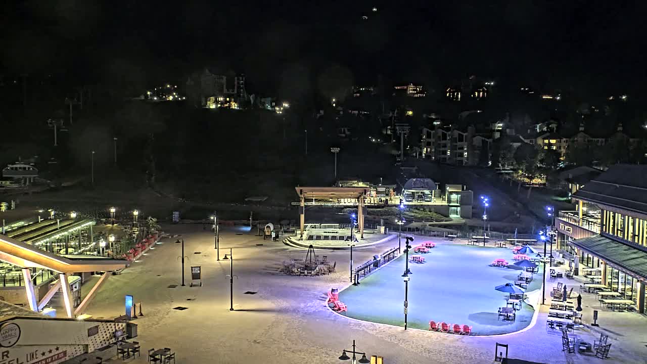 A nighttime aerial view of a snow-covered plaza with buildings and outdoor seating areas, illuminated by artificial lights, nestled against a hillside of residential buildings.
