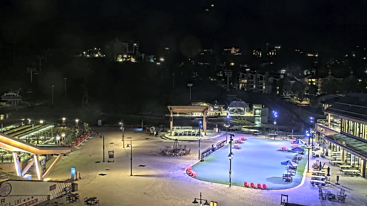 An outdoor plaza at night, illuminated by lights, features a skating rink, buildings, and seating areas, under a clear, dark sky.