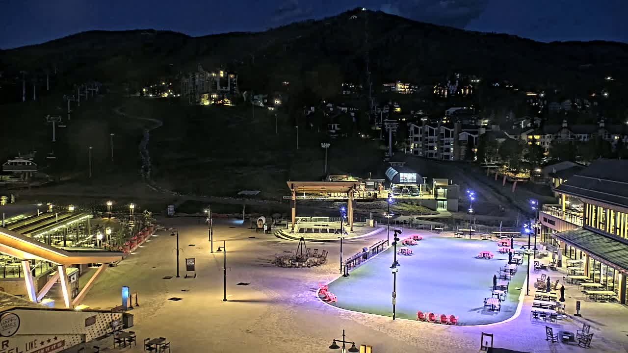A nighttime view of an illuminated outdoor plaza with a skating rink, surrounded by buildings and ski lifts on a mountainside under a dark sky.