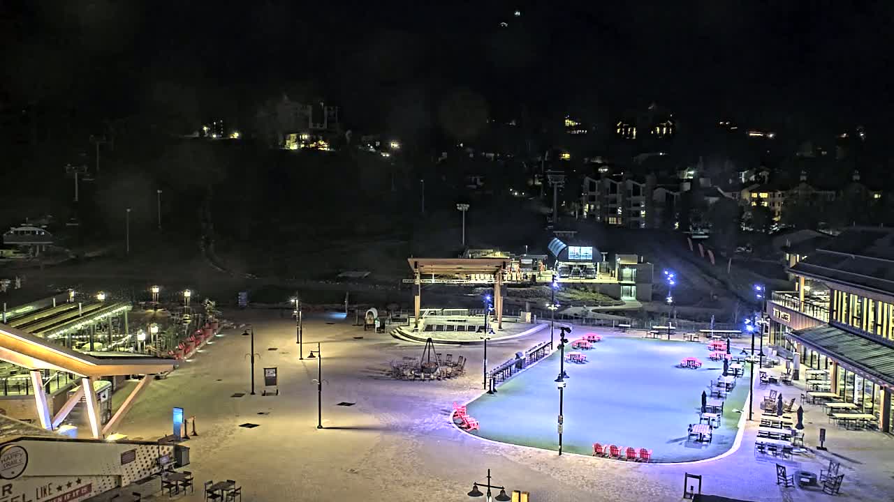 A snow-covered plaza at night features a lit-up ice rink surrounded by outdoor seating and buildings, with hills and residences visible in the background under a clear, dark sky.
