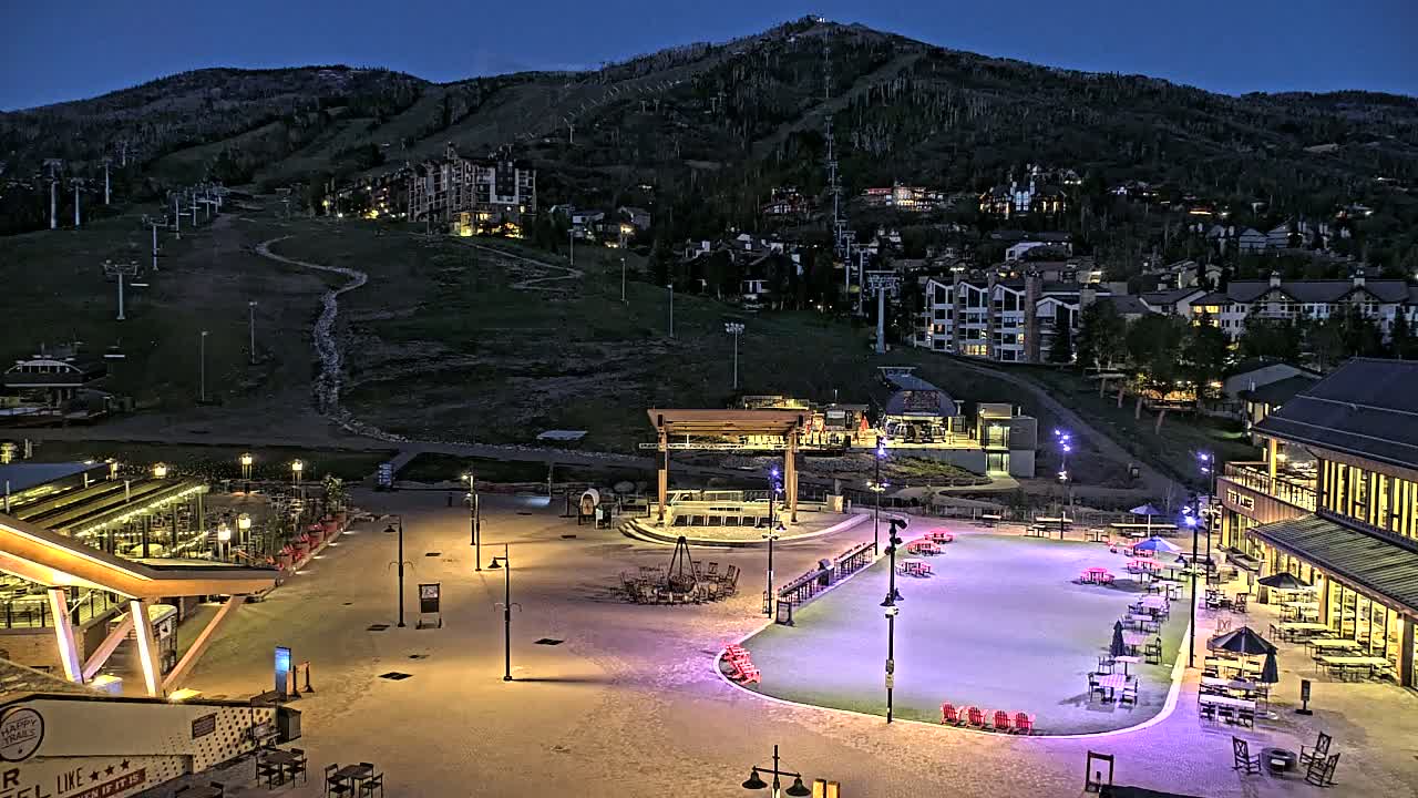 A brightly lit plaza with outdoor seating and a skating rink sits at the base of a mountain at night, with ski lifts visible on the mountainside.