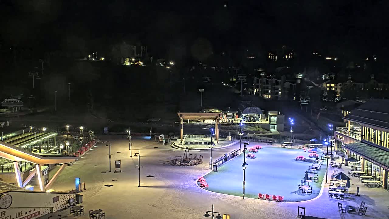 An outdoor ice rink at night, surrounded by snow-covered ground, buildings, and outdoor seating areas, is illuminated under a dark sky.