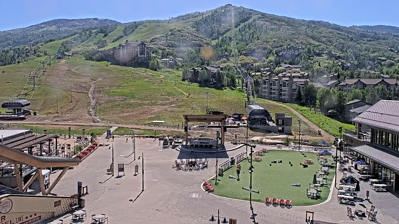 An outdoor plaza with seating areas, a stage, and a grassy patch sits below a ski lift and mountainside buildings on a sunny day.