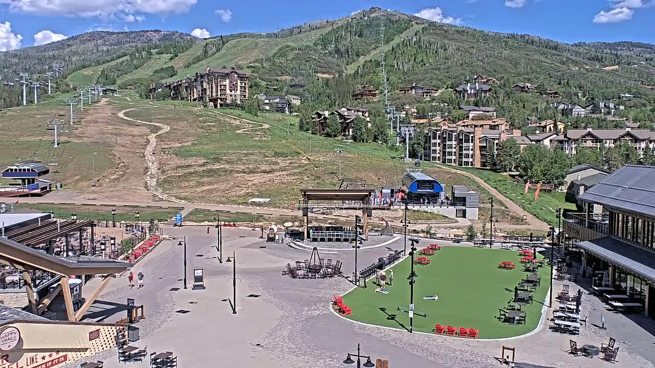 A sunny day at a mountain resort shows an outdoor plaza with seating areas, a stage, and a ski lift in the background, all situated amidst buildings and a grassy hillside.