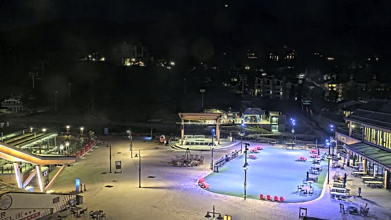 A snow-covered plaza at night with outdoor seating and a brightly lit ice rink, overlooked by buildings and a hillside with homes.